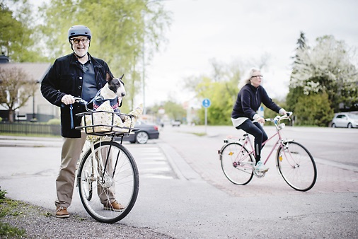 En glad man med cykelhjälm på huvudet som leder cykel och kvinna som cyklar i bakgrunden i en gatukorsning en solig dag.