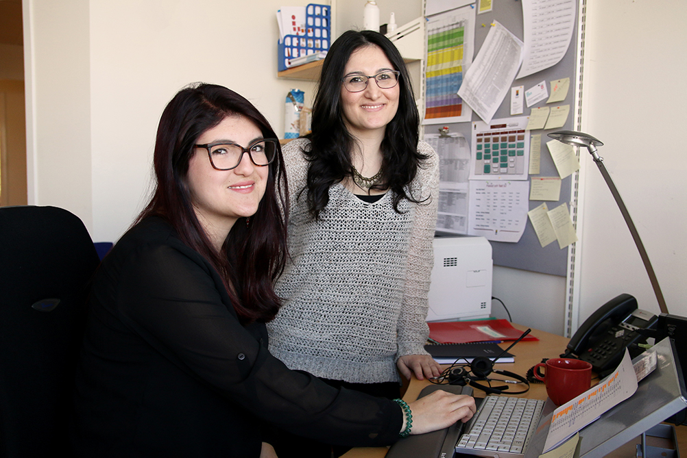 Hülya Yesil och Valentina Restrepo Pulido sitter och står vid ett skrivbord på ett kontor.