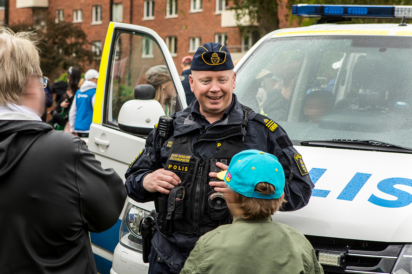 En polis står vid en polisbil och pratar med ett barn och en vuxen, ser glad ut.
