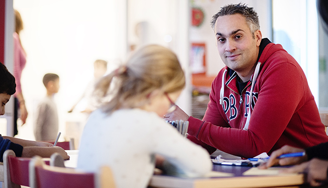 En lärare tittar mot kameran sittandes i ett klassrum där barn sitter och arbetar vid sina bänkar.