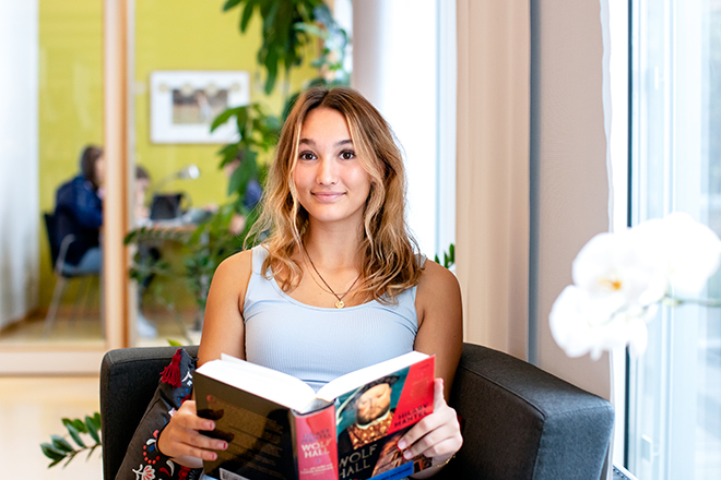 Tjej i gymnasieåldern som sitter i skolans bibliotek med en bok och ler mot kameran.