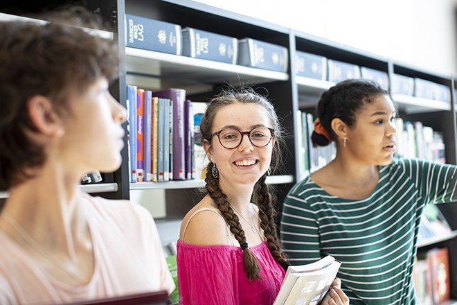 Tre tjejer som står framför en bokhylla i skolans bibliotek varav en tittar rakt in i kameran och ler.