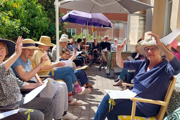 Några äldre personer sitter på en altan i solen, vinkar och tittar in i kameran.